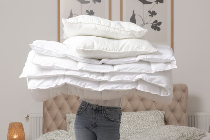A person holds a large stack of white pillows and bedding in a bedroom with a tufted headboard and floral wall art, reminiscent of the fresh linens after expert wedding dress cleaning Syracuse.
