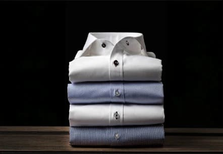 Four neatly folded dress shirts, two white on top and two light blue underneath, stacked on a wooden surface against a black background.