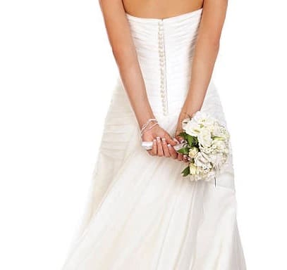 A woman in a strapless white wedding dress holds a bouquet of white flowers behind her back.