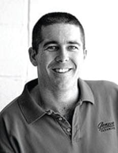 A man with short dark hair is smiling and wearing a collared shirt. The photo is black and white and taken indoors against a light background.