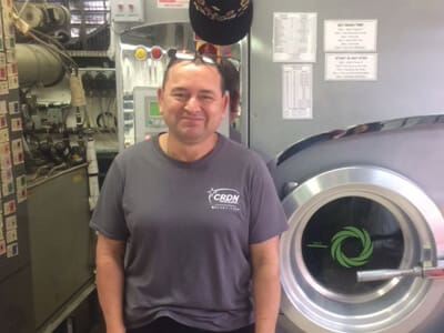 A man in a gray CRDN t-shirt stands in front of industrial laundry machines inside a laundromat.