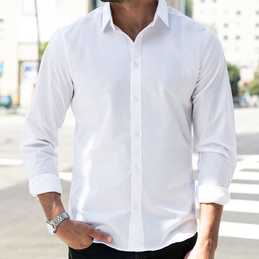 A man wearing a white button-down shirt with rolled-up sleeves stands outdoors with one hand in his pocket.