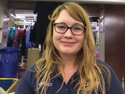 A woman with long, wavy blonde hair and glasses is standing indoors, wearing a navy blue shirt with a name tag. Laundry and clothing racks are visible in the background.