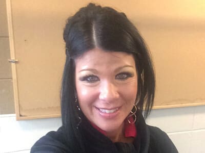 A woman with long black hair and hoop earrings smiles at the camera in front of a beige bulletin board.