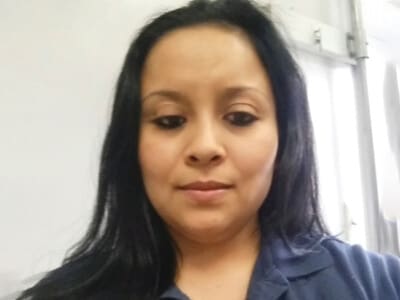 A woman with long black hair wears a navy blue collared shirt and looks down slightly indoors, with a light-colored background and window behind her.