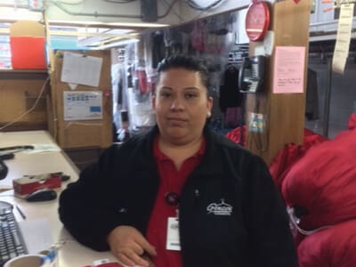 A woman in a work uniform stands behind a counter in a dry cleaning shop, with red laundry bags and equipment visible in the background.