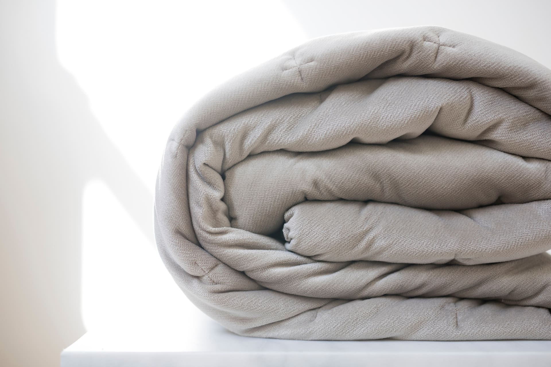 A neatly rolled, light beige blanket sits on a white surface with sunlight casting a shadow in the background.