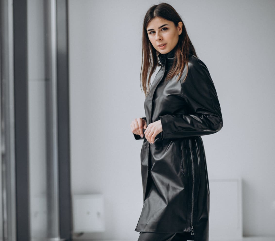 A woman stands indoors wearing a long black leather coat, looking towards the camera with her hands near the coat's buttons.