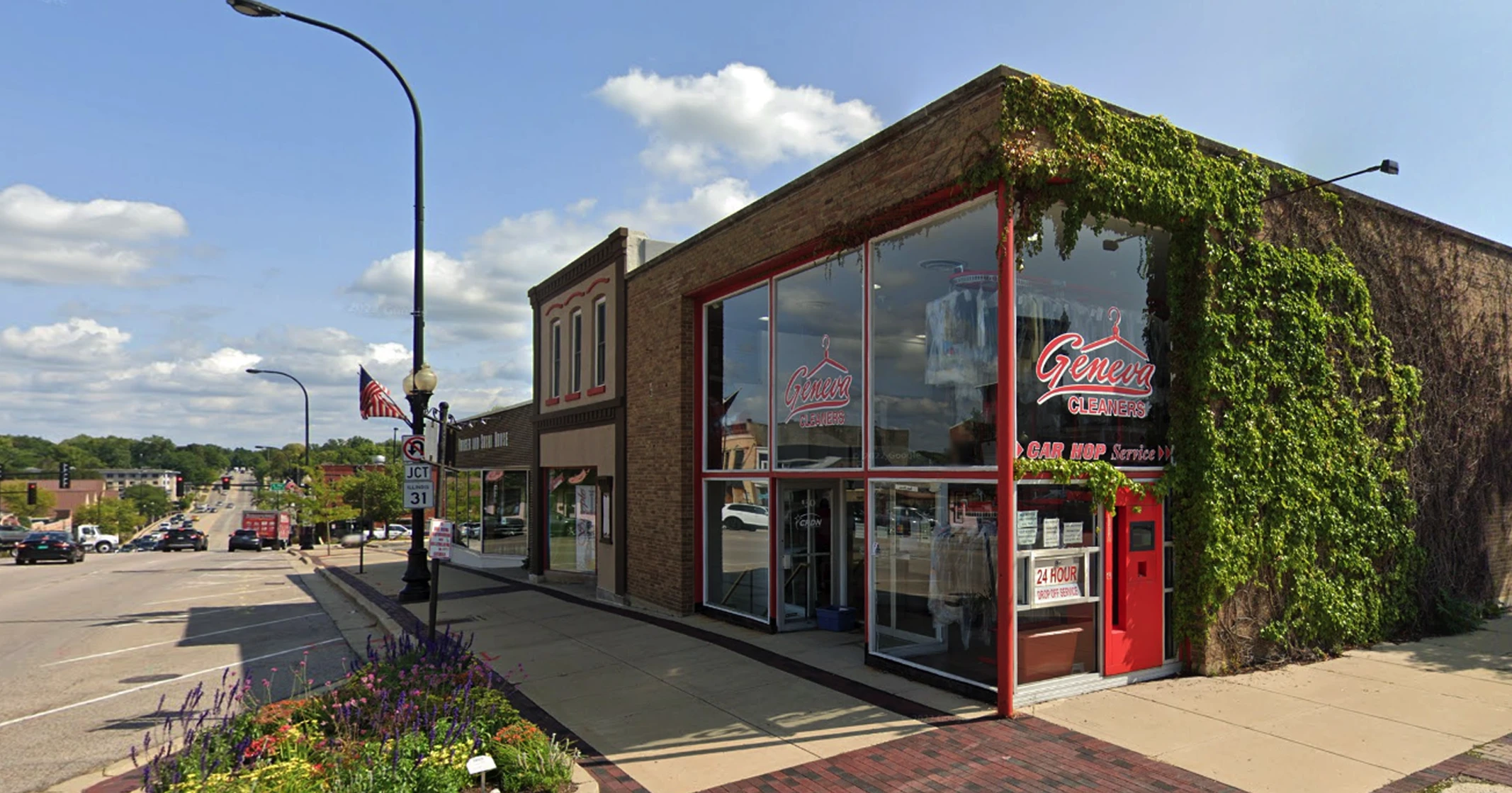 Street view of a brick building with large windows and red trim, housing a business called Geneva Cleaners; sidewalk, street, and other shops are visible under a partly cloudy sky.