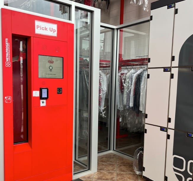 Red kiosk labeled "Pick Up" next to lockers and a glass room filled with hanging clothes in a dry cleaning or laundry facility.