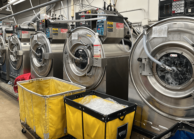 Four industrial washing machines are running in a laundry facility, with two large yellow laundry carts positioned in front of them.