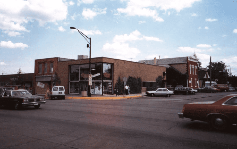 A brick corner building with large windows sits at an intersection, with parked and moving cars nearby under a partly cloudy sky.