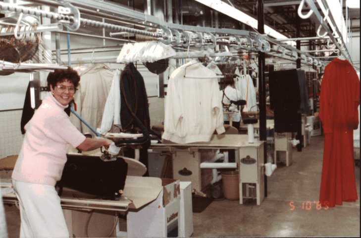 A woman uses an iron at a dry cleaning facility, with clothing hanging on racks and garment presses in the background.