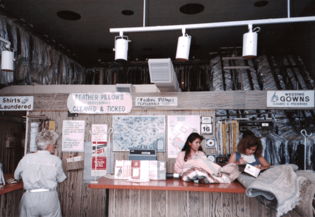 Three people stand at a dry cleaning counter with racks of clothes in plastic behind them, and signs advertising pillow cleaning and wedding gown preservation are visible above the counter.