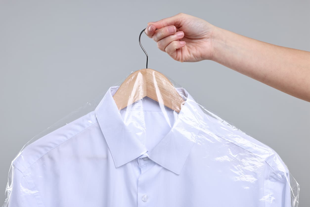 A hand holds a hanger with a neatly pressed white dress shirt covered in clear plastic, typically used for dry cleaning.