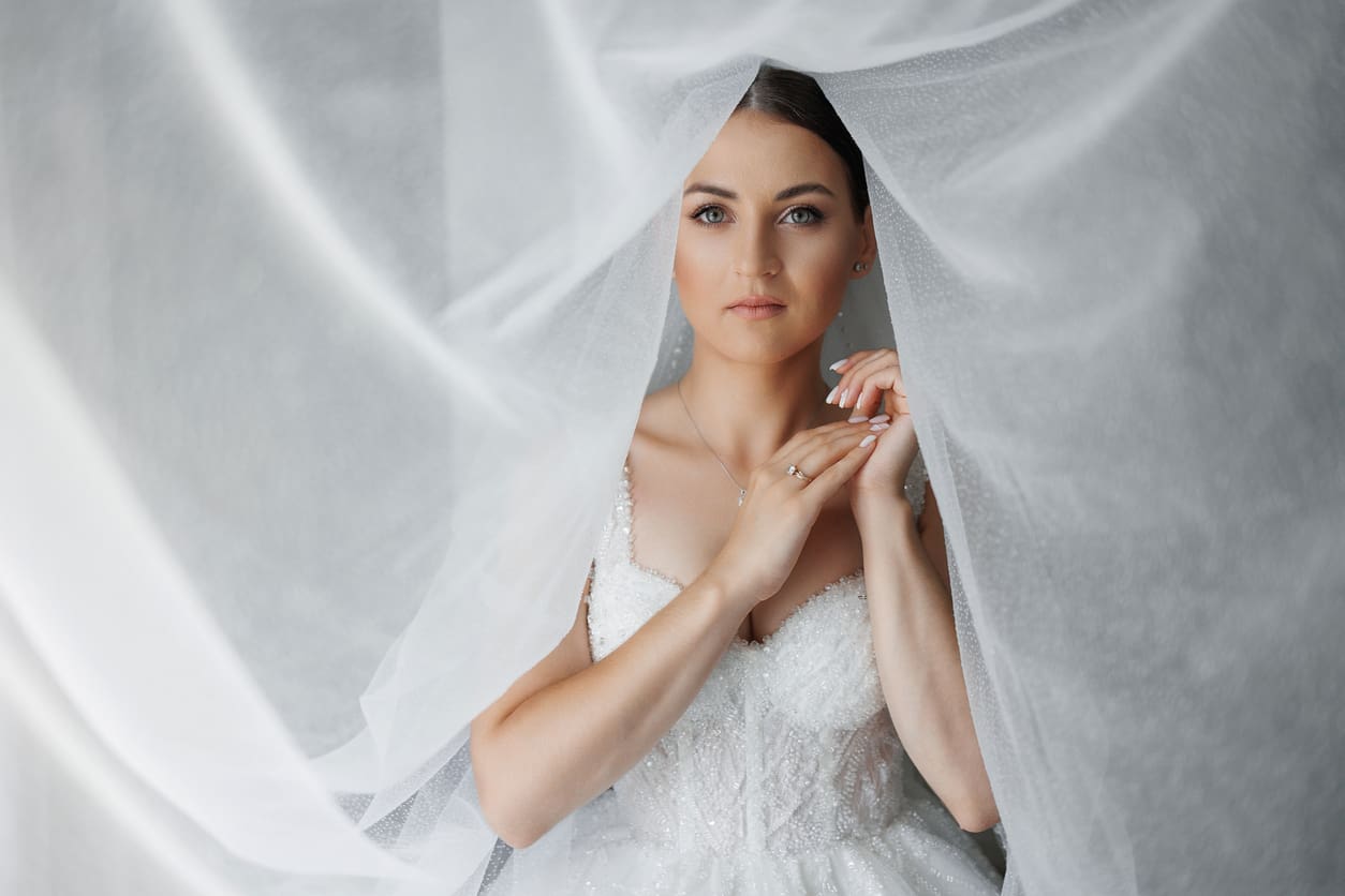 A woman in a white wedding dress stands under a sheer veil, looking directly at the camera with her hands gently touching her chest.