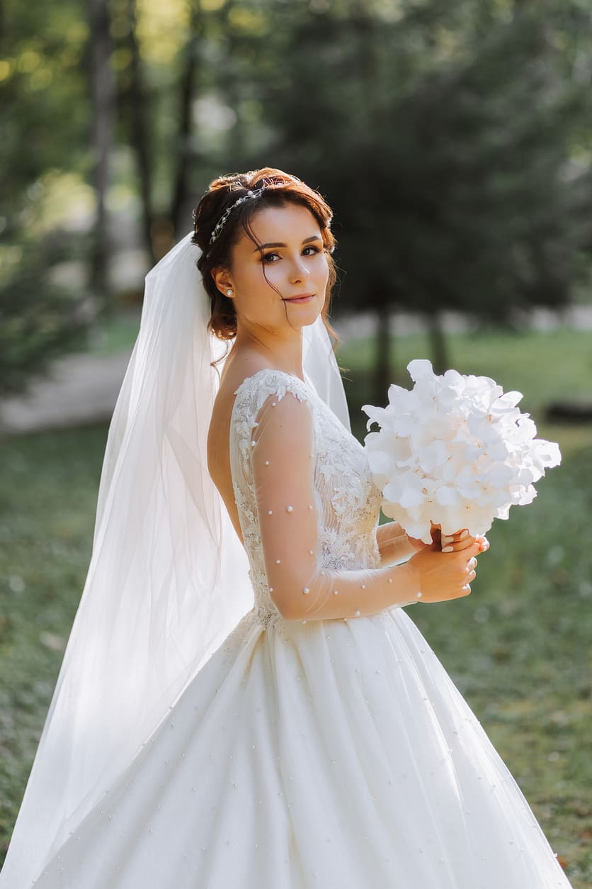A bride in a white wedding dress and veil holds a bouquet of white flowers while standing outdoors in a green, sunlit area.