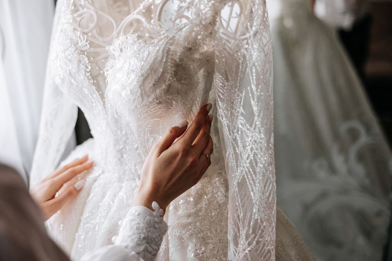 Close-up of hands adjusting a detailed, white lace wedding dress with intricate beadwork and embroidery.