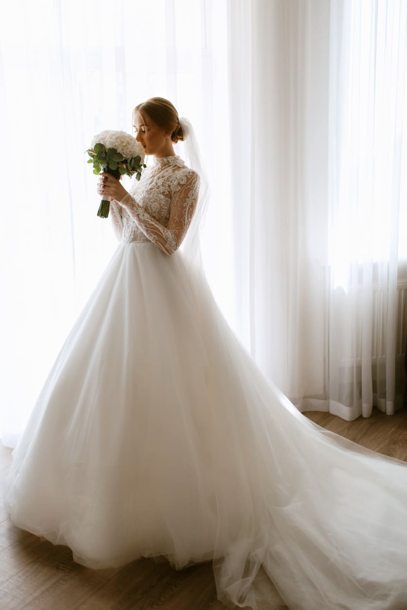A bride in a white long-sleeve lace wedding dress holds a bouquet of flowers and stands by a window with sheer curtains.