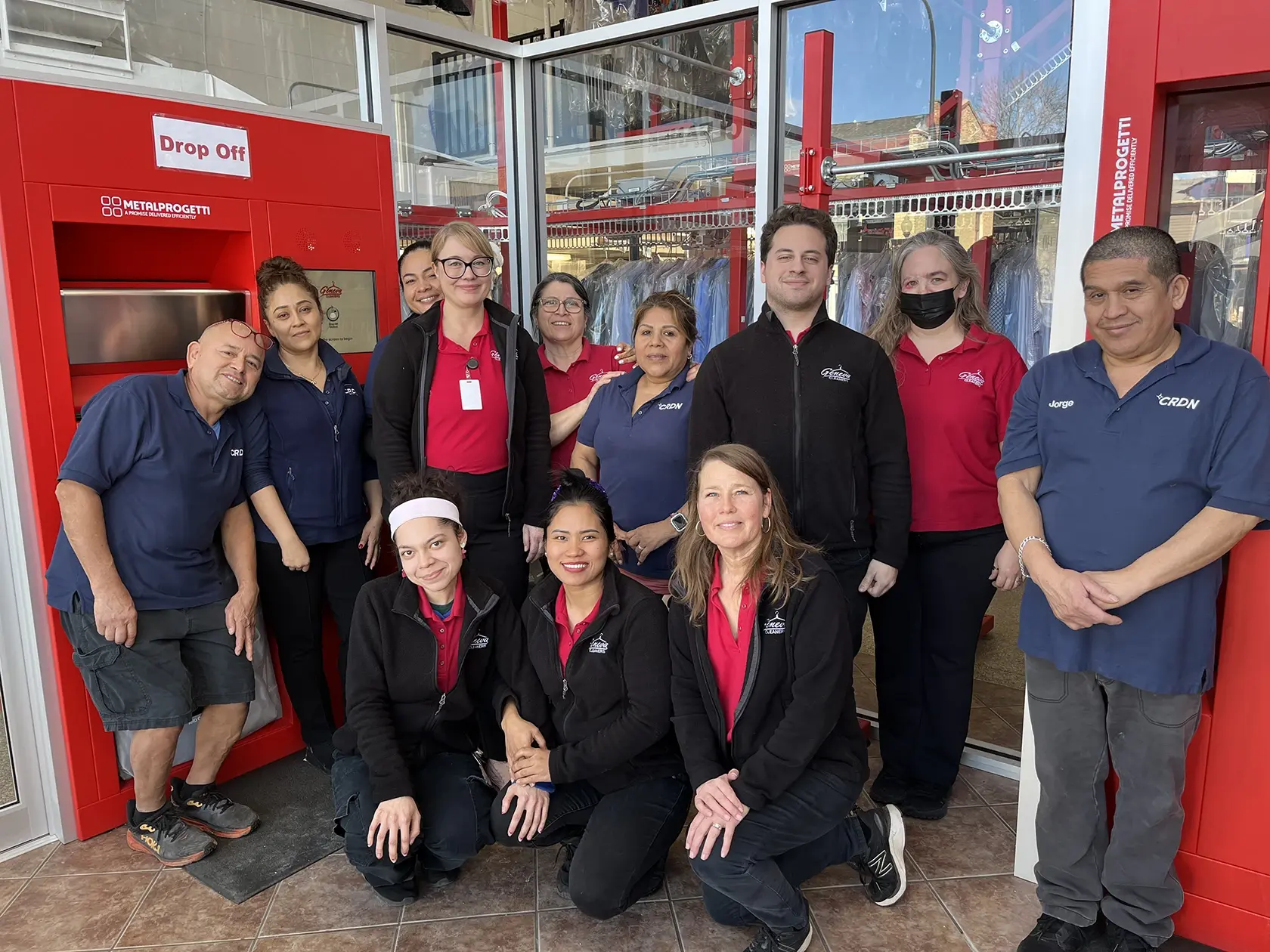 A group of eleven employees pose together in front of a dry cleaning business counter, with red and blue uniforms and a drop-off kiosk visible in the background.