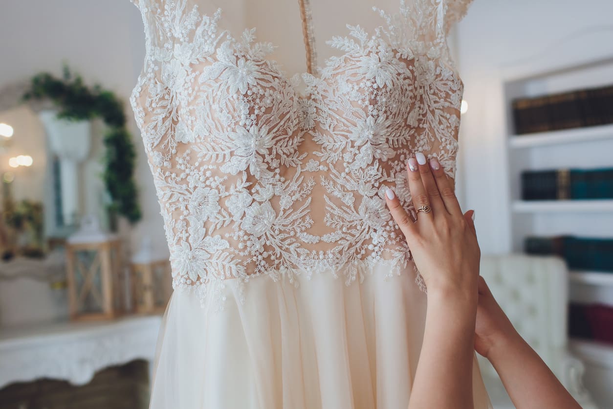 A person with manicured nails touches the detailed lace bodice of an ivory wedding dress hanging in a well-lit room.