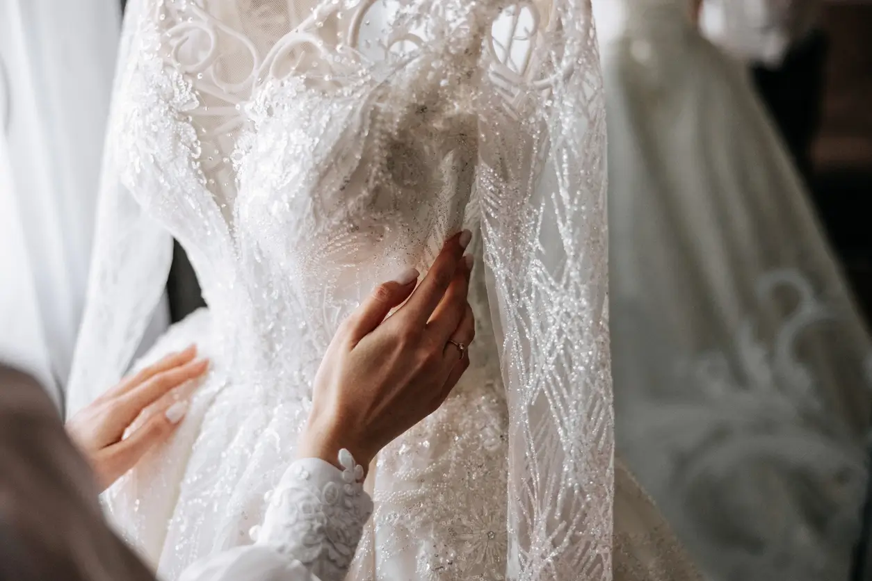 A person&rsquo;s hands touching the intricate beadwork and lace of a long-sleeved white wedding gown.