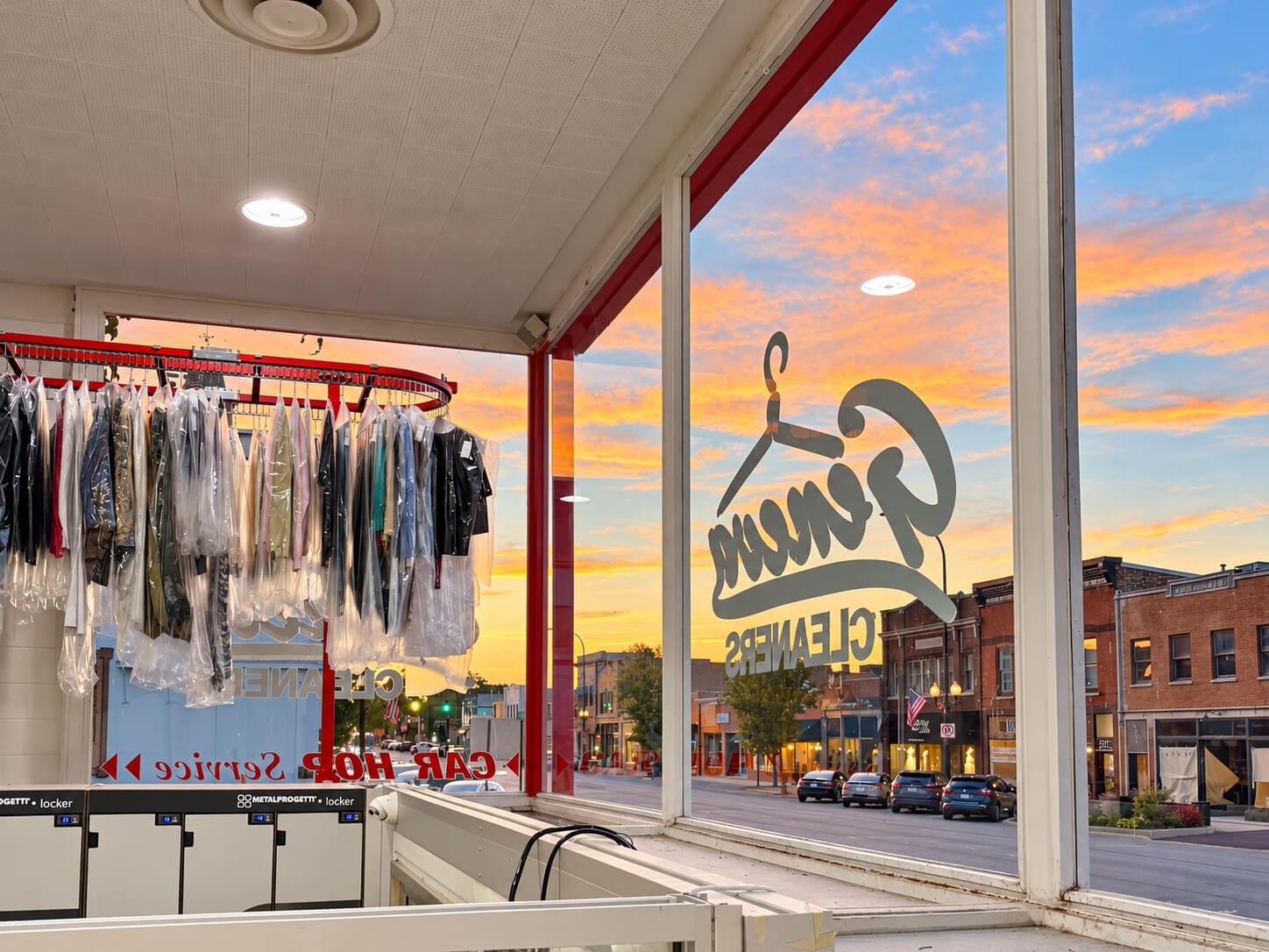 View from inside a dry cleaner looking out at a sunset over a street with cars and buildings.