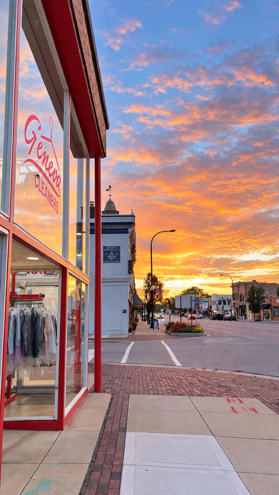 Street view of a small town during a vibrant orange and pink sunset, showing the storefront of Geneva Cleaners.